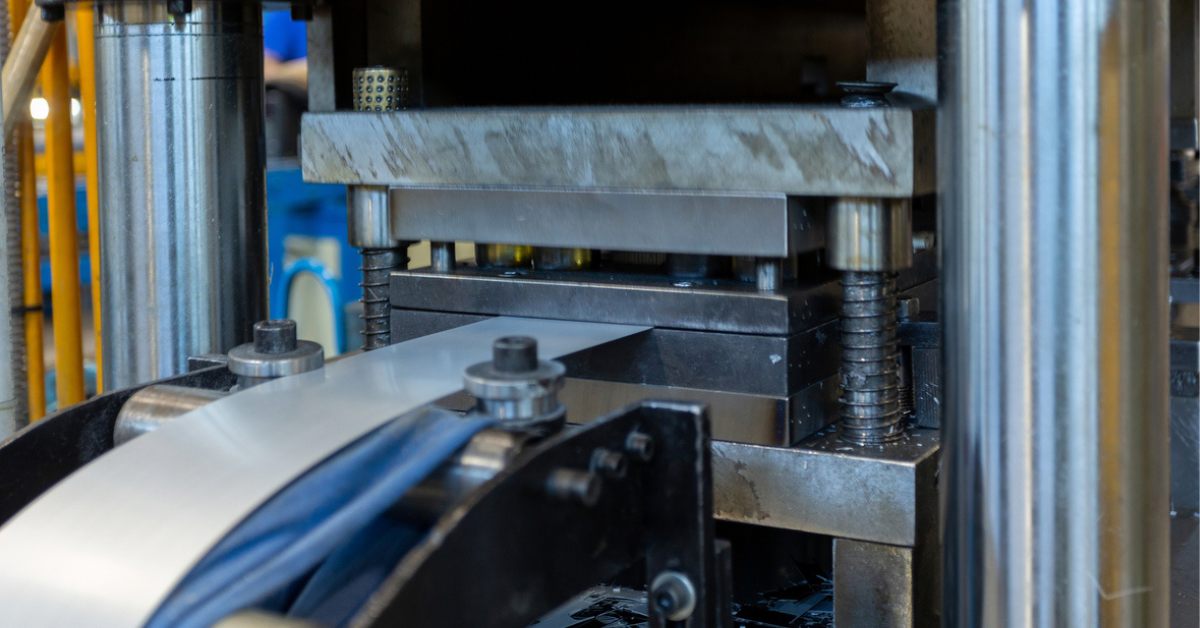 Close-up of an industrial press with spring-loaded columns compressing a rectangular die as a blue conveyor belt feeds a strip of sheet metal into the machine.